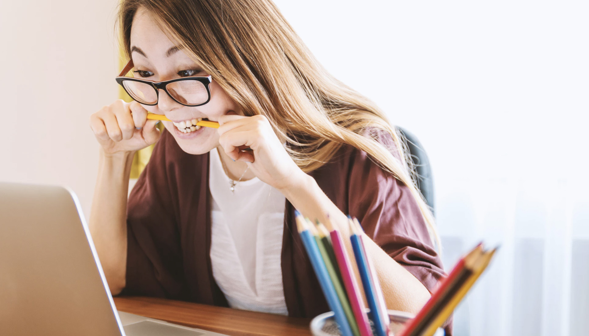 A girl chewing her pencil while looking at the computer because she is stressed